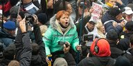 Eine Frau in grüner Jacke steht in einer Menschenmenge und ruft etwas