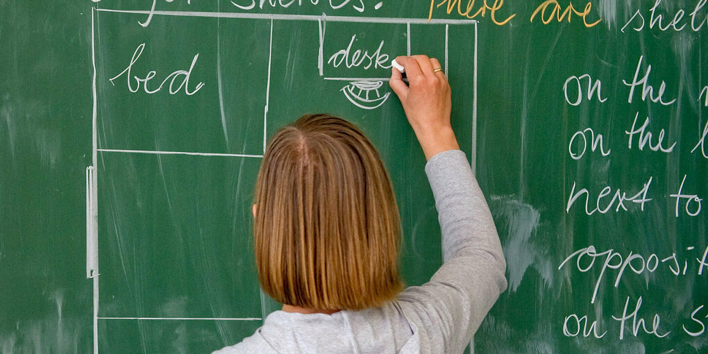 Man sieht eine Person mit blondem langem Haar von hinten. Sie steht an einer Schultafel und schreibt darauf etwas mit Kreide