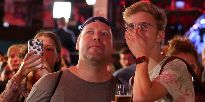 Zwei Männer schauen auf einen Bildschirm, einer hält sich eine Hand vor den Mund