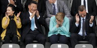Merkel und Xi Jinping sitzen in einem Fußballstadion. Xi Jinping klatscht und schaut lachend zu Merkel, diese beugt sich nach vorne und versteckt ihr Gesicht in ihren Händen. Sie wirkt so, als würde sie gerade sehr lachen