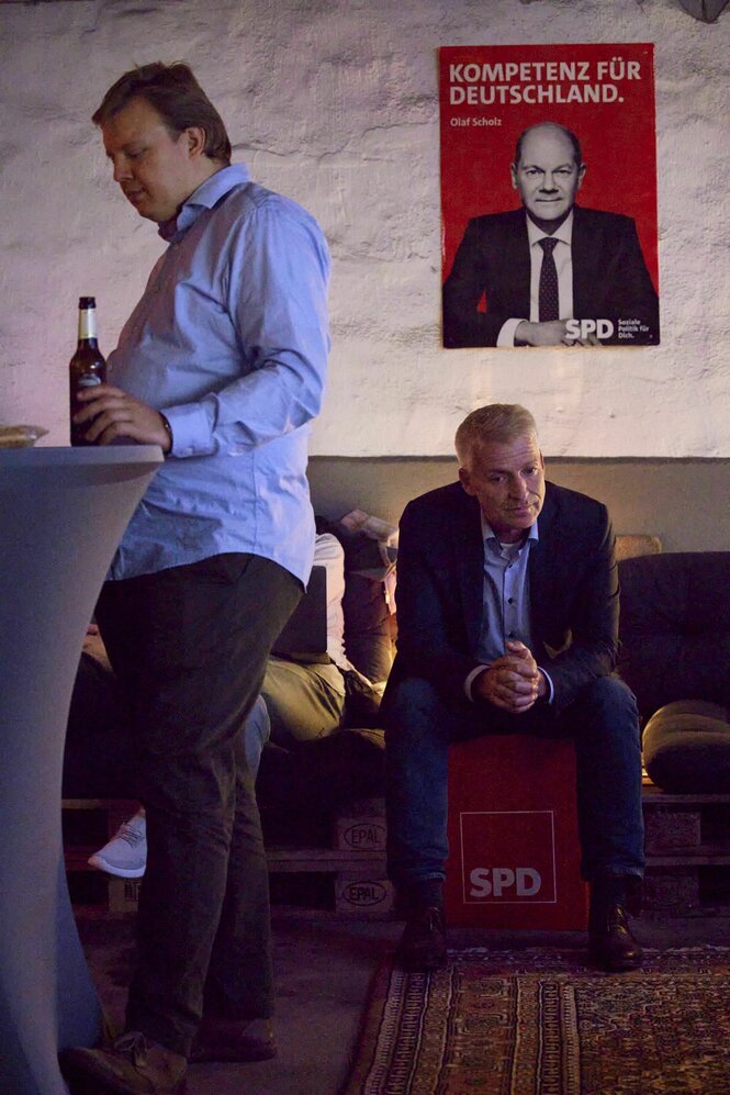 Ein Mann sitzt auf einem Sofa, neben ihm steht ein Mann mit einer Flssche Bier am Stehtisch und an der Wand ein Plakat von Olaf Scholz