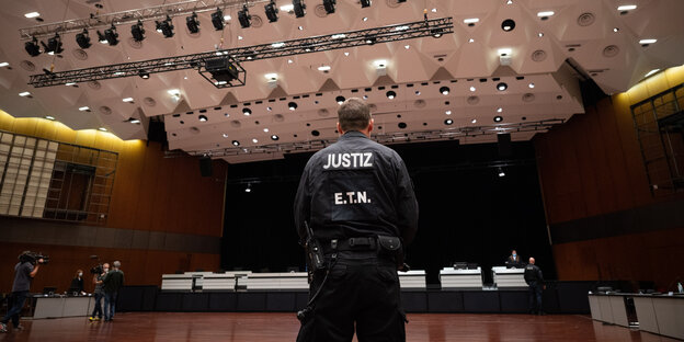 Mann mit "Polizei"-Aufschrift auf Jacke steht mit dem Rücken zum Betrachter vor leeren Justizsaal