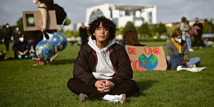 Der Fridays for Future Aktivist Quang Paasch sitzt auf einer Wiese vor dem Bundestag. Hinter ihm ist ein Schild zu erkennen mit der Aufschrift "Die Uhr tickt". Ganz hinten hebt sich die Silhouette des Kanzler:innenamt ab.