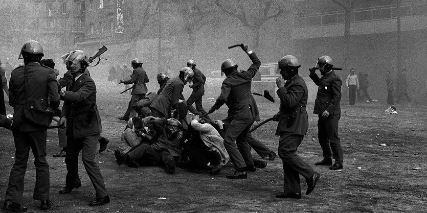 Schwarz/Weiß-Aufnahme, düstere Stimmung, Polizisten mit weißen Helmen schlagen auf am Boden liegenden Demonstranten ein