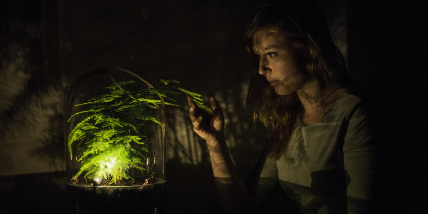 Eine junge Frau in einem dunklen Raum, der nur von einer Lampe neben einer Art Farn erleuchtet wird