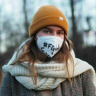 Eine junge Frau mit mit gelber Mütze, einem weißen Mund-Nasen-Schutz, auf dem in schwarzer Schrift etwas nicht ganz Erkennbares geschriebensteht und dickem Schal und Jacke. Eine junge Frau mit mit gelber Mütze, einem weißen Mund-Nasen-Schutz, auf dem in schwarzer Schrift etwas nicht ganz Erkennbares geschriebensteht und dickem Schal und Jacke.