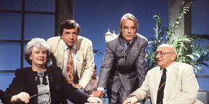 Eine Frau und drei Männer in Anzügen in einem Fernsehstudio.