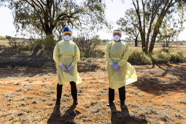 zwei Personen sind komplett in Mülläcken und Schutzplastik mit Mundschutz und so weiter gekleidet. sie stehen auf sandigem Boden, dahinter Bäume. Die Sonne scheint und es sieht sehr warm aus