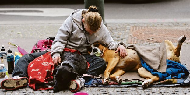 Eine Frau sitzt mit ihren Habseligkeiten und zwei Hunden auf einer Straße.
