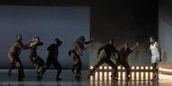 sechs Tänzer in dunkler Kleidung tanzen vor dem Sänger in weißer Unterwäsche