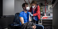 Ein junger Mann mit Bart und Maske sitzt in einem alten Bremer Linienbus, der als Impfmobil dient. Die Frau, die ihn impfen wird, hält die Spritze in der Hand und begutachtet seinen linken Oberarm. Rechts im Bus steht ein Tisch mit Desinfektionsmittel