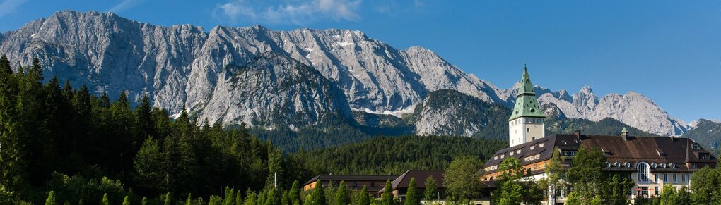 Ein Blick auf das Schlosshotel Elmau mit dem Wettersteingebirge im Hintergrund bei fast wolkenlosem blauen Himmel.