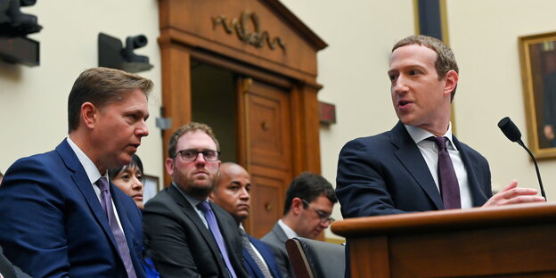 Mark Zuckerberg steht an einem Pult und dreht sich zu einer Reihe von Männern im Anzug hinter ihm um