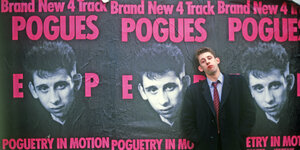 Der Musiker Shane MacGowan steht vor einer Wand, die mt Postern der Band "Pogues" beklebt ist