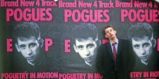 Der Musiker Shane MacGowan steht vor einer Wand, die mt Postern der Band "Pogues" beklebt ist