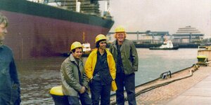 Historisches Foto aus den 70ern. Vor einem Hafenbecken stehen drei Arbeiter der AG Weser aus Bremen mit gelben Helmen. Sie lächeln. Ein vierter Mann schaut zu ihnen herüber. Im Hintergrund ein großes Schiff