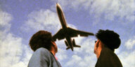 Zwei Frauen schauen in den Himmel, über ihnen fliegt direkt ein Flugzeug