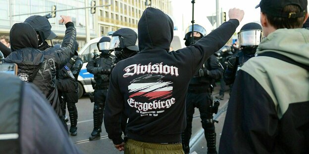 Ein Mann trägt einen schwarzen Hoodie mit der Aufschrift "Division Erzgebirge" reckt seine Faust gegenüber Polizisten in die Höhe
