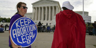 Eine Frau mit einem runden "Keep Abortion Legal"-Plakat und eine Frau von hinten mit rotem Umhang vor dem Supreme Court in Washington