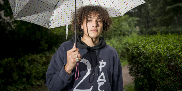 Ein junger Mann mit Locken und einem 2 Pac-Pullover hält einen gepunkteten Regenschirm