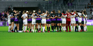 Fußballspielerinnen beider Teams bilden auf dem Rasen einen Kreis