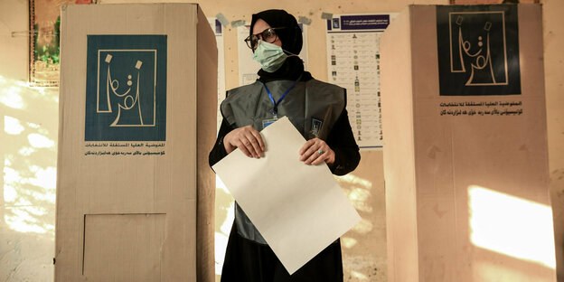 Eine Person hält ein großes Blatt Papier und steht zwischen zwei Wahlkabinen