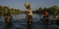 Fluechtlinge überqueren einen Fluß, ein Kind, das auf den Schultern eines Mannes getragen wird, hebt sein Stoffspielzeug in die Höhe