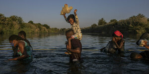 Fluechtlinge überqueren einen Fluß, ein Kind, das auf den Schultern eines Mannes getragen wird, hebt sein Stoffspielzeug in die Höhe