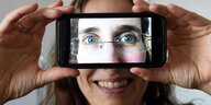 Eine Frau hält ein Handy vor ihre Augen, auf dem Handydisplay sind die Augen der Frau zu sehen