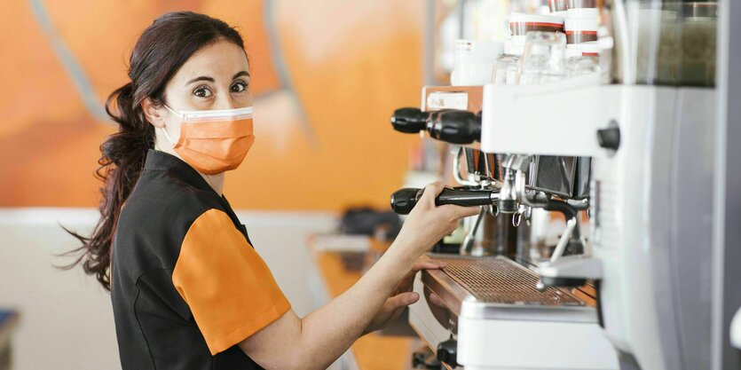 Junge Frau mit Gesichtsmaske steht an der Kaffeemaschine in einem Cafe