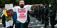 Ein Mann mit Basecap und einem T-Shirt mit der Aufschrift: Stop Genocid on Cameroonians - bewacht von Polizisten