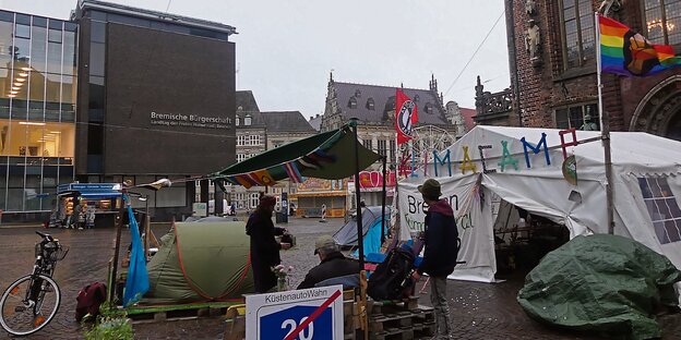 Das Klimacamp in Bremen; drei Aktivist:innen stehen unter einem kleinen Vorzelt, hinter ihnen das große Gemeinschaftszelt mit Antifa- und Regenbogenflagge. Im Hintergrund Rathaus und Bürgerschaft sowie Karussels für den bald startenden Freimarkt