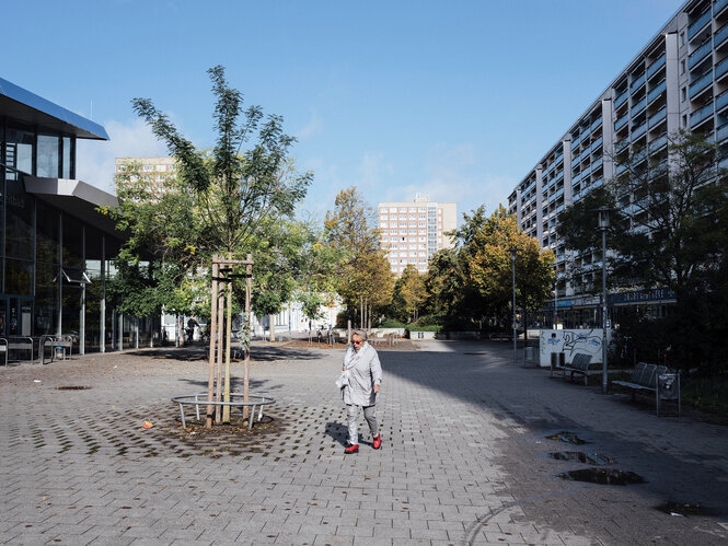 ÄltereFrau geht über eine menschenleere Straße, gesäumt von Plattenbauten