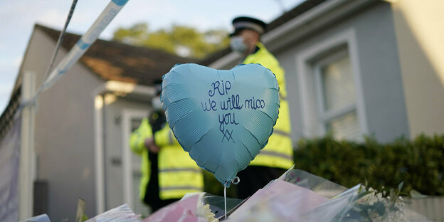 Ein Herz-Luftballon mit der Aufschrift RIP We will miss you" schwebt über auf der Erde abgelegte Blumensträuße. Im Hintergrund Polizisten vor einem Haus