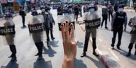 Eine Hand zeigt den Driefinger-Gruß, im Hintergrund sind Polizisten mit Schutzschilden zu sehen