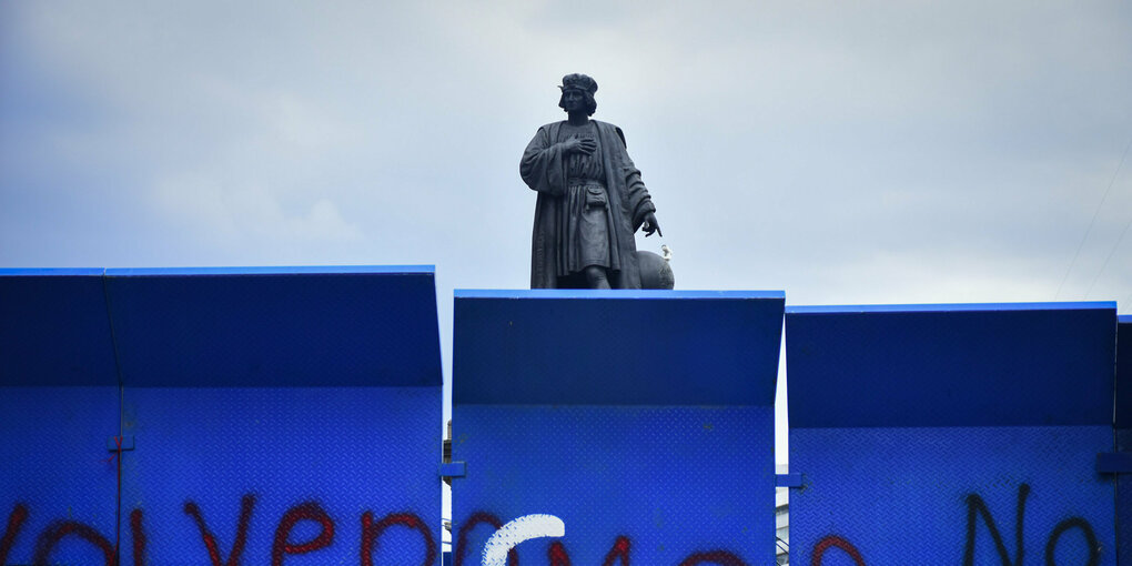 Hinter einem Bauzaun steht eine Statue mit dem für Kolumbus typischen Barett