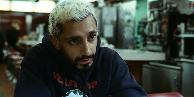 Schauspieler Riz Ahmed als "Ruben" im Filmdrama „Sound of Metal“. Er trägt einen schwarzen Bart und hat weiß gefärbte Haare. Hinter ihm sind Barstühle und eine Theke verschwommen abgebildet