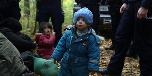Ein kleines Kind aus dem Irak mit Mütze wird von Polizisten im Wald umringt