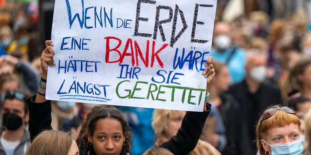 Auf einer Demonstration hält eine Aktivistin ein Schild hoch mit dem Text "Wenn die Erde eine Bank wäre hättet ihr sie längst gerettet"