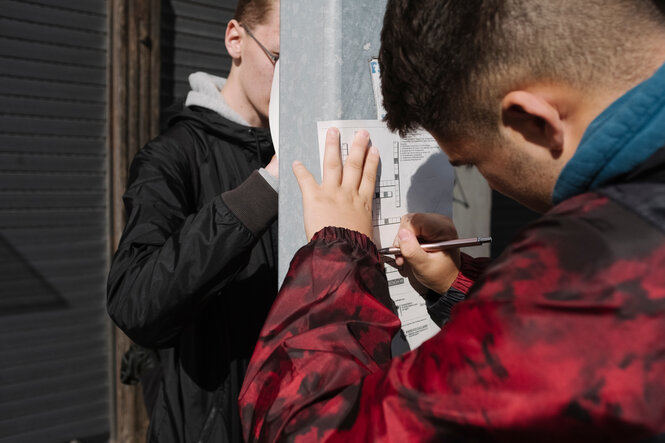 Jugendliche lösen ein Kreuzworträtsel, sie drücken die Zettel an die Wand, weil sie keinen Tisch haben