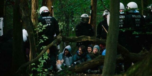 Umwelt-Aktivist:innen sitzen im Hambacher Forst auf dem Boden und sind umringt von Polizist:innen in voller Montur