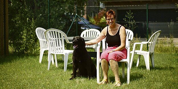 Eine Frau sitzt auf einem weißen Plastikstuhl in einem Garten und krault ihren Hund.