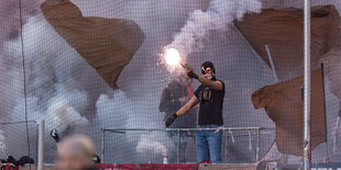 Ein Ultra des FC St. Pauli in schwarzer Vermummung hält Pyrotechnik vor einem Fanblock mit zwei schwarzen fahnen, die komplett eingenebelt sind