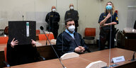 Die Angeklagte im Gerichtssaal neben ihrem Verteidiger, hält sich einen Aktenordner vors Gesicht, im Hintergrund drei PolizistInnen