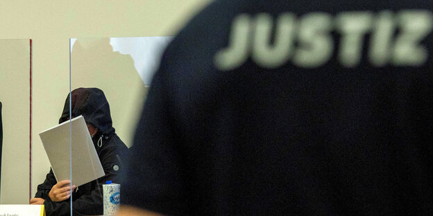 Eine Person hält eine Mappe vor ihr Gesicht, davor eine Jacke mit der Aufschrift Justiz