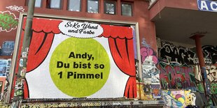 An einer Hauswand hängt ein Plakat mit roten Vorhängen, die aufgezogen sind. In der Mitte steht: "Andy, du bist so 1 Pimmel", darüber steht: "Soko Wand und Farbe"