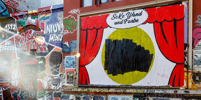 An einer Hauswand hängt ein Plakat mit roten Vorhängen, die aufgezogen sind.Die Botschaft in der Mitte ist mit schwarz übermalt, darüber steht: "Soko Wand und Farbe"