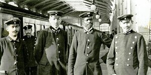 Vier Männer stehen 1902 in Bahnuniformen vor einem Zug. Das Bild zeigt den afrodeutschen Aktivist Martin Dibobe (2. von rechts) und Hochbahn-Kollegen