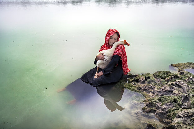 Eine Frau in Kleidung, unter anderem mit Kopftuch, sitzt auf einem Felsen im Wasser. Das Wasser reicht ihr bis zur Hüfte. In ihren Armen hält sie einen großen Vogel.