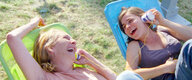 Zwei Frauen liegen auf einer Wiese, lachen und halten sich ein Dosentelefon an die Ohren